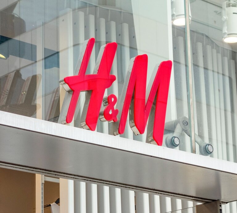 Das Bild zeigt das leuchtend rote Logo von H&M, das an einer Ladenfront befestigt ist. Das Logo ist vor einem gläsernen Hintergrund mit Sicht auf innenliegende Strukturen und Gegenstände platziert. Darüber sind moderne Beleuchtungselemente angebracht. | © Adobe Stock/  IB Photography