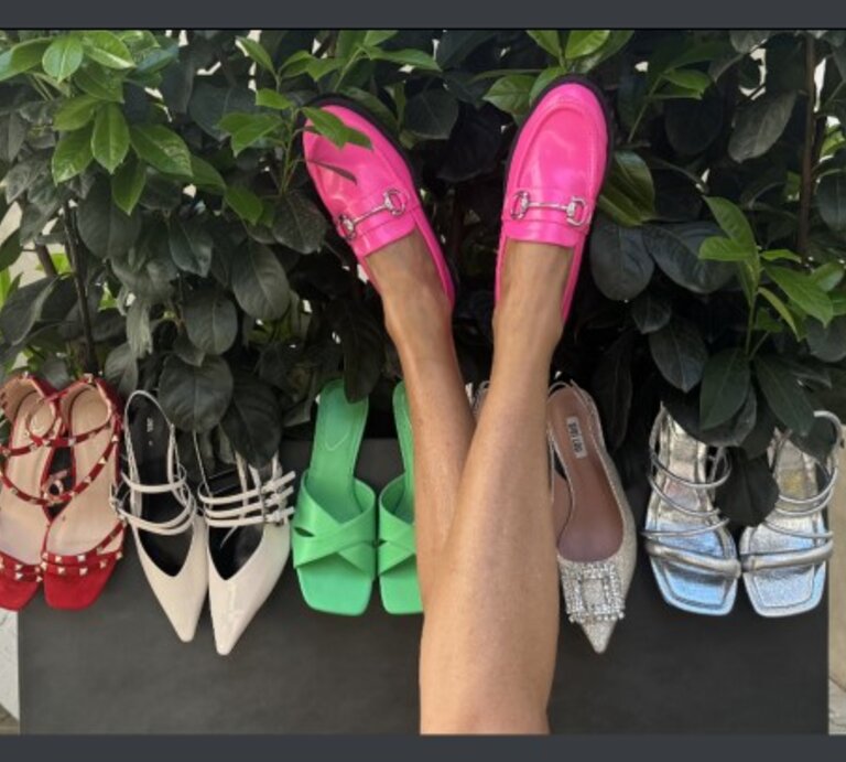 Auswahl an Damenschuhen, die vor einer grünen Pflanze arrangiert sind. In der Mitte des Bildes sind die Beine einer Person zu sehen, die leuchtend pinke Loafer trägt. Die Schuhe, die um die Pflanze herum aufgestellt sind, umfassen verschiedene Stile und Farben. | © Renate Zott