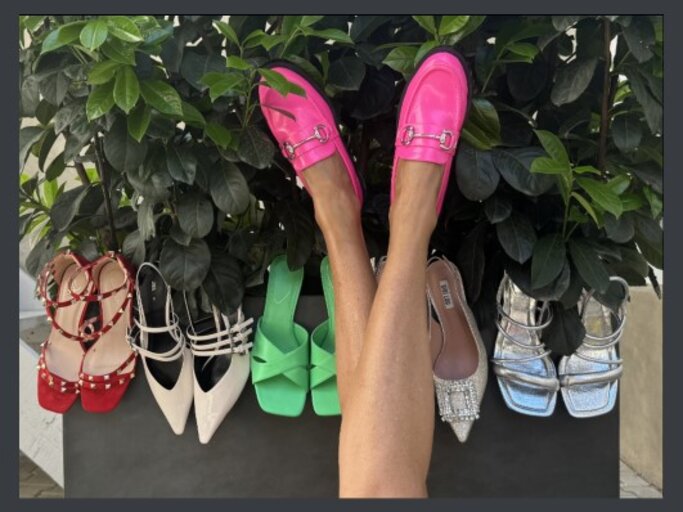 Auswahl an Damenschuhen, die vor einer grünen Pflanze arrangiert sind. In der Mitte des Bildes sind die Beine einer Person zu sehen, die leuchtend pinke Loafer trägt. Die Schuhe, die um die Pflanze herum aufgestellt sind, umfassen verschiedene Stile und Farben. | © Renate Zott