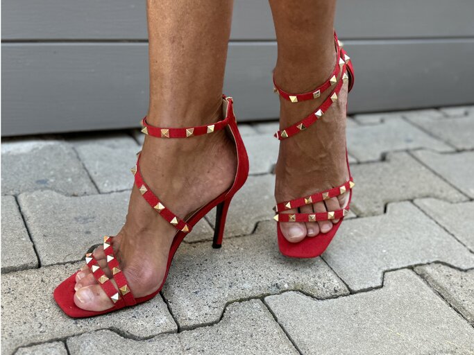 Beine, die in roten High Heels mit goldenen Nieten stecken. Diese Schuhe haben mehrere dünne Riemen, die den Fuß und den Knöchel umschließen, und einen hohen Stilettoabsatz. Die goldenen Nieten sind gleichmäßig auf den Riemen verteilt und verleihen den Schuhen einen auffälligen und stilvollen Look. | © Renate Zott