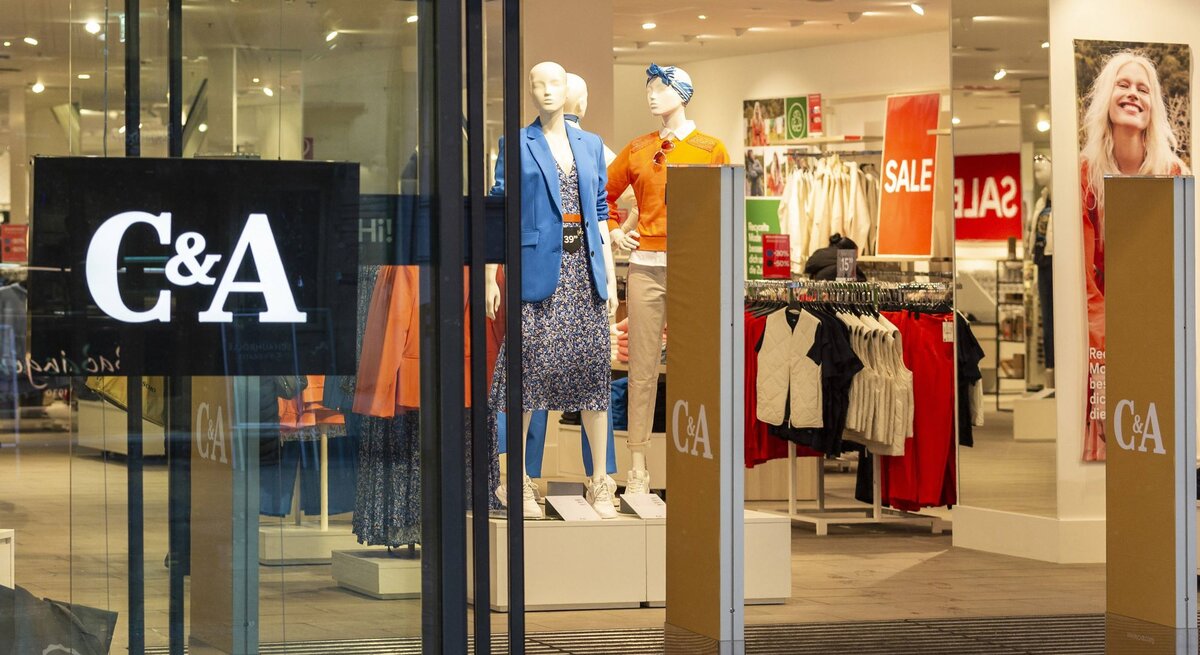  Das Foto zeigt den Eingang eines C&A-Geschäfts mit großen Glasscheiben, durch die man in den Innenraum blicken kann. Zwei Mannequins sind im Schaufenster ausgestellt, eines trägt eine blaue Jacke und das andere eine orange Bluse. Innerhalb des Ladens sind SALE-Schilder sichtbar, die auf Rabatte hinweisen. Auf der Glasfront ist das C&A-Logo deutlich zu sehen. Die Szene wirkt einladend und ist typisch für Einzelhandelsgeschäfte in einer Einkaufsstraße.
