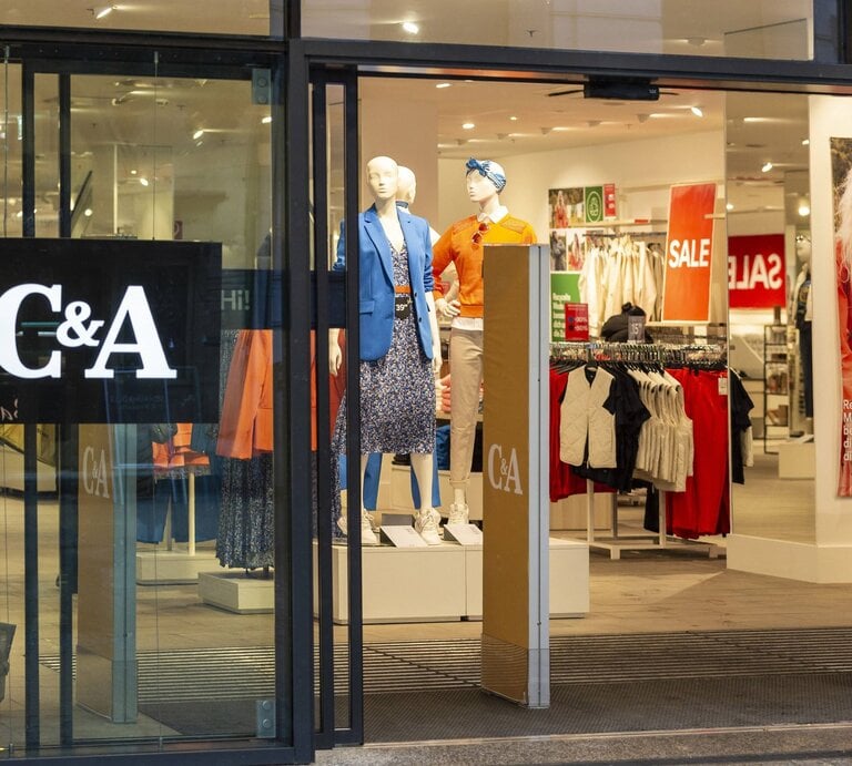  Das Foto zeigt den Eingang eines C&A-Geschäfts mit großen Glasscheiben, durch die man in den Innenraum blicken kann. Zwei Mannequins sind im Schaufenster ausgestellt, eines trägt eine blaue Jacke und das andere eine orange Bluse. Innerhalb des Ladens sind SALE-Schilder sichtbar, die auf Rabatte hinweisen. Auf der Glasfront ist das C&A-Logo deutlich zu sehen. Die Szene wirkt einladend und ist typisch für Einzelhandelsgeschäfte in einer Einkaufsstraße. | © IMAGO / CHROMORANGE