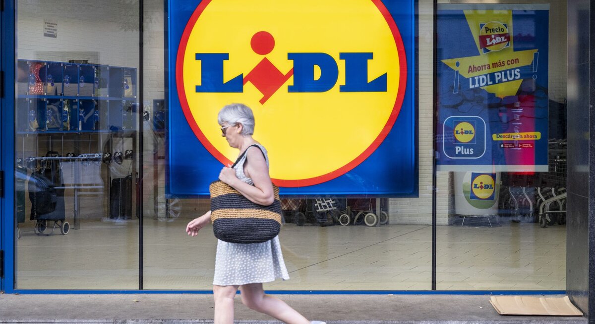 Grauhaarige Frau in weißem Sommerkleid und Sneakers läuft an einer Lidl-Filiale mit großem gelbem Logo vorbei.