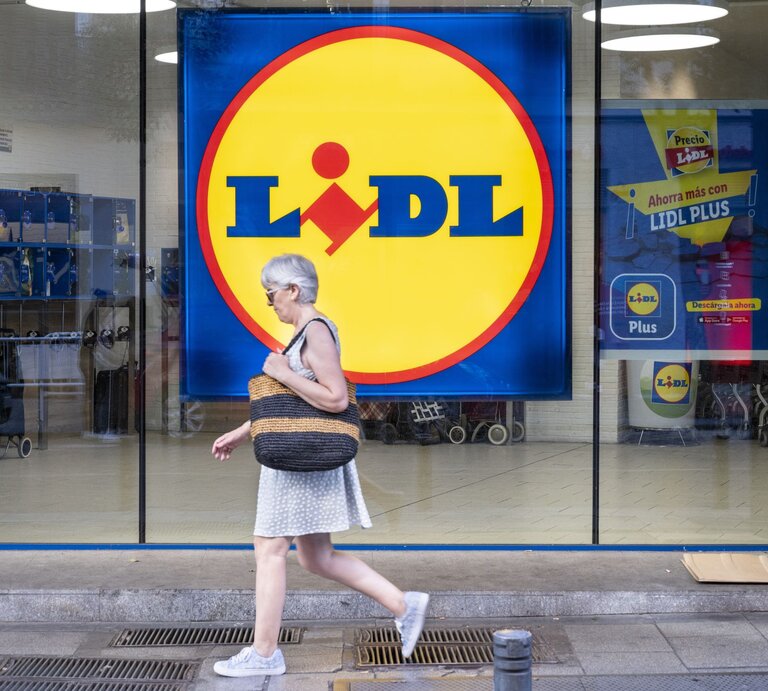 Grauhaarige Frau in weißem Sommerkleid und Sneakers läuft an einer Lidl-Filiale mit großem gelbem Logo vorbei. | © Xavi Lopez/SOPA Images/LightRocket via Getty Images