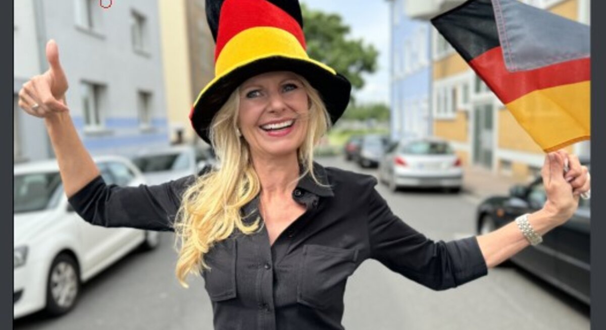 Frau, die eine große, farbenfrohe Hut in den Farben der deutschen Flagge trägt und eine Deutschlandflagge in der Hand hält. Sie lächelt und zeigt einen Daumen hoch, während sie auf einer Straße steht.