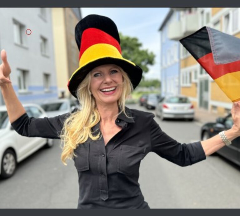 Frau, die eine große, farbenfrohe Hut in den Farben der deutschen Flagge trägt und eine Deutschlandflagge in der Hand hält. Sie lächelt und zeigt einen Daumen hoch, während sie auf einer Straße steht. | © Renate Zott