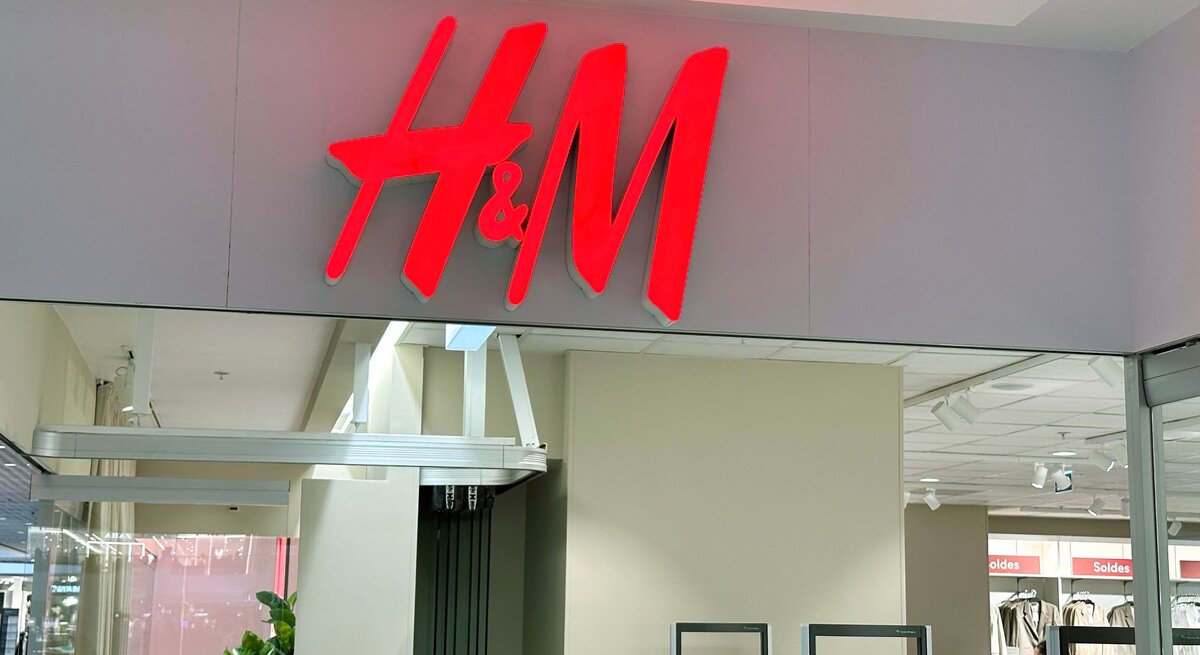 Leuchtschild des H&M-Logos in Rot über dem Eingang einer Filiale mit sichtbarem Innenraum und Kleidungsständern.