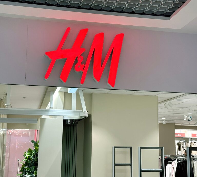 Leuchtschild des H&M-Logos in Rot über dem Eingang einer Filiale mit sichtbarem Innenraum und Kleidungsständern. | © Adobe Stock/ IB Photography