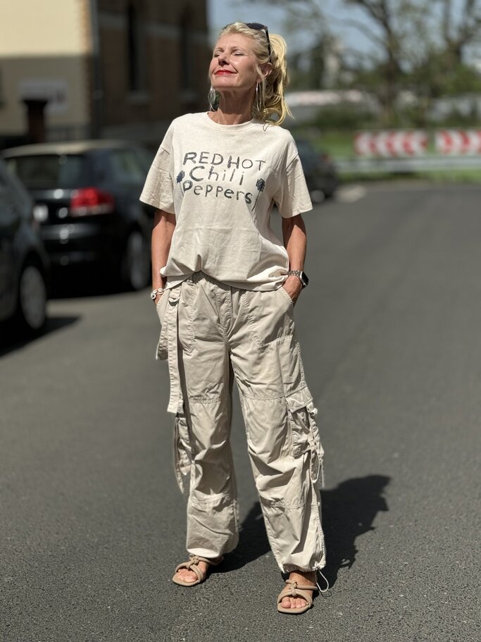 Frau, die ein oversized T-Shirt mit der Aufschrift "Red Hot Chili Peppers" trägt, kombiniert mit hellen Fallschirmhosen. Sie trägt dazu flache Sandalen und eine Sonnenbrille, die auf ihrem Kopf ruht. | © Renate Zott