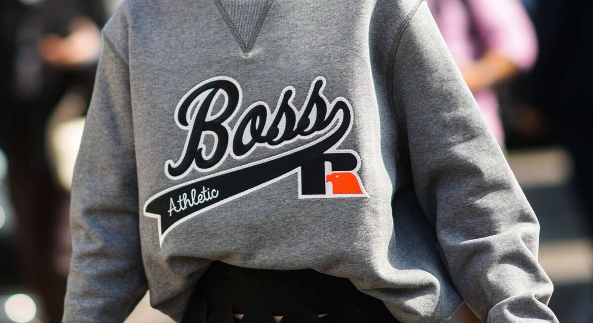 Nahaufnahme einer Frau, die ein graues Sweatshirt trägt. Auf der Vorderseite des Sweatshirts ist in Großbuchstaben das Wort "Boss" zusammen mit dem Zusatz "Athletic" aufgedruckt. Ein markantes Detail ist das eingenähte V-förmige Dreieck am Kragen, das typisch für viele Sweatshirt-Designs ist.