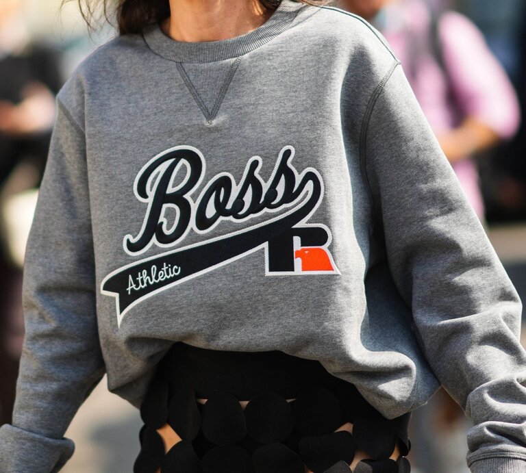 Nahaufnahme einer Frau, die ein graues Sweatshirt trägt. Auf der Vorderseite des Sweatshirts ist in Großbuchstaben das Wort "Boss" zusammen mit dem Zusatz "Athletic" aufgedruckt. Ein markantes Detail ist das eingenähte V-förmige Dreieck am Kragen, das typisch für viele Sweatshirt-Designs ist. | © Getty Images / Edward Berthelot