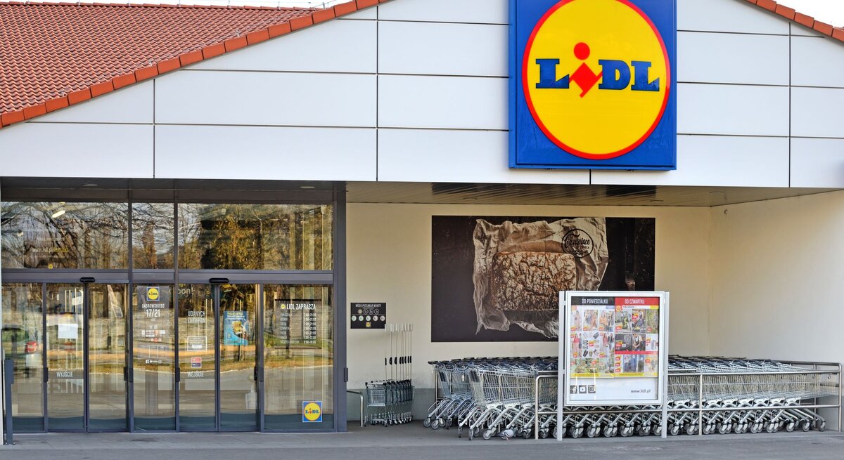 Auf dem Foto ist der Eingangsbereich einer Lidl-Filiale zu sehen. Über dem Eingang prangt das charakteristische Firmenschild mit gelbem Kreis und dem Lidl-Logo in der Mitte. Das Gebäude hat ein schlichtes Design mit einer weißen Fassade und einem roten Ziegeldach. Vor dem Eingang sind Glasautomatiktüren und eine Reihe von Einkaufswagen. An der linken Seite des Eingangs befindet sich ein Plakat mit Werbung für Produkte, die im Laden erhältlich sind. Im Hintergrund sind Bäume und ein parkähnlicher Bereich zu erkennen.