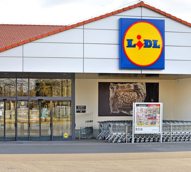 Auf dem Foto ist der Eingangsbereich einer Lidl-Filiale zu sehen. Über dem Eingang prangt das charakteristische Firmenschild mit gelbem Kreis und dem Lidl-Logo in der Mitte. Das Gebäude hat ein schlichtes Design mit einer weißen Fassade und einem roten Ziegeldach. Vor dem Eingang sind Glasautomatiktüren und eine Reihe von Einkaufswagen. An der linken Seite des Eingangs befindet sich ein Plakat mit Werbung für Produkte, die im Laden erhältlich sind. Im Hintergrund sind Bäume und ein parkähnlicher Bereich zu erkennen. | © Adobe Stock/ Tomasz Warszewski