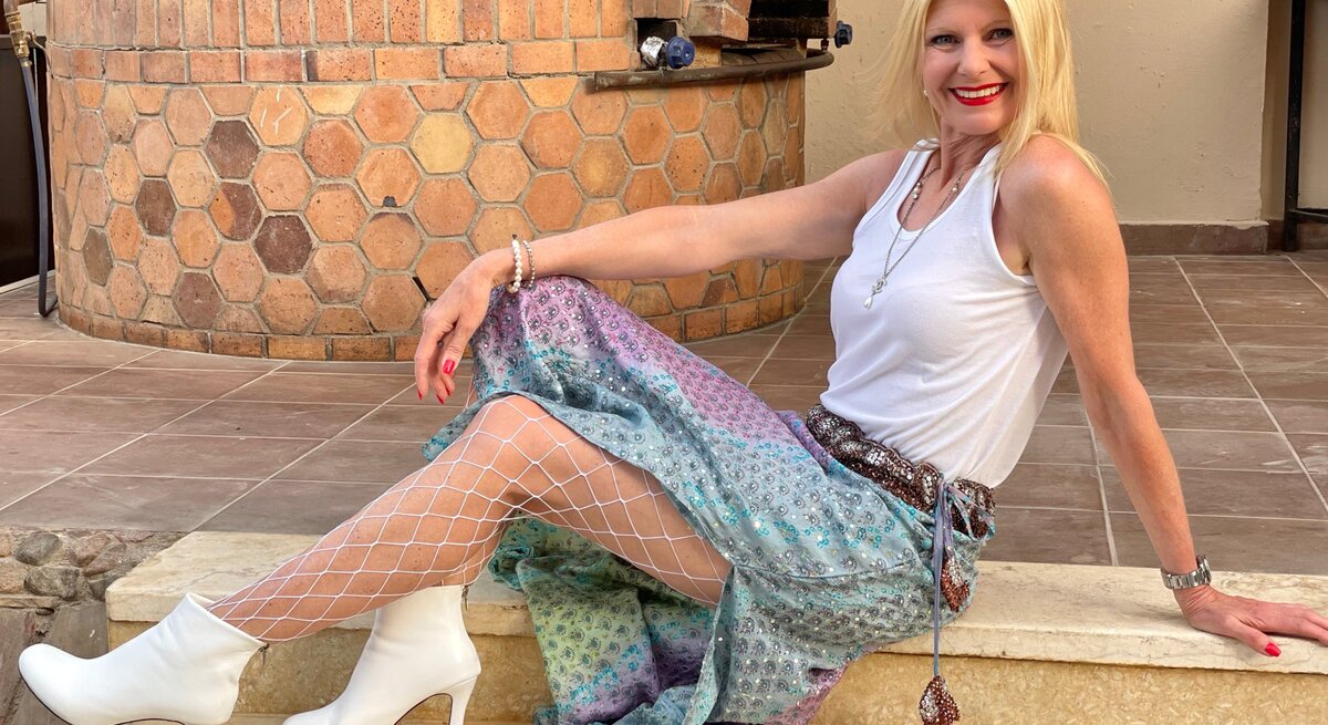 Renate Zott sitzt lächelnd auf einer steinernen Treppe und hat ein lebhaftes Outfit an. Sie hat blonde Haare, trägt ein weißes Tanktop und darüber eine bunte, schimmernde Hose mit einer Netzstrumpfhose darunter und weiße Stiefeletten.