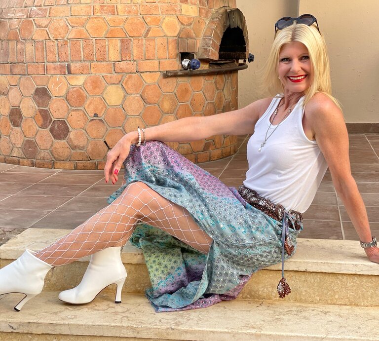 Renate Zott sitzt lächelnd auf einer steinernen Treppe und hat ein lebhaftes Outfit an. Sie hat blonde Haare, trägt ein weißes Tanktop und darüber eine bunte, schimmernde Hose mit einer Netzstrumpfhose darunter und weiße Stiefeletten. | © Renate Zott
