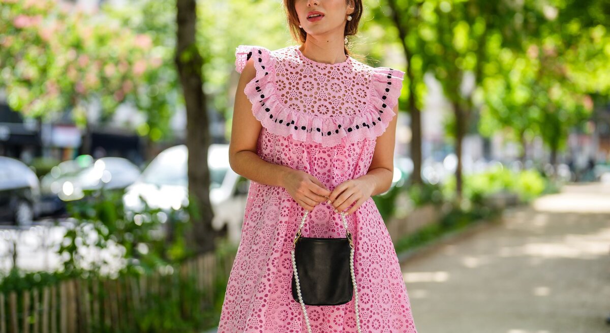 Elegante Frau in einem leuchtend pinken Sommerkleid mit Lochstickerei, unterwegs auf einem städtischen Gehweg.