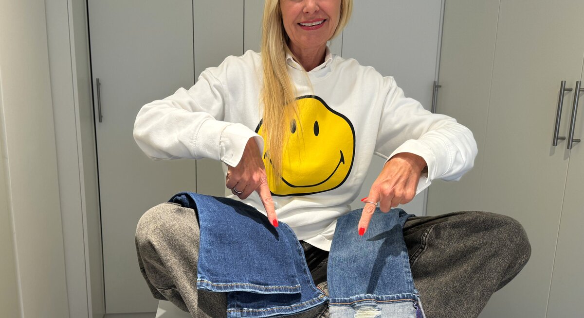 Renate Zott trägt lächelnd einen weißen Pullover mit einem großen, gelben Smiley-Gesicht darauf und zeigt auf zwei verschiedene Jeansstoff-Muster, die sie auf ihren Knien platziert hat.