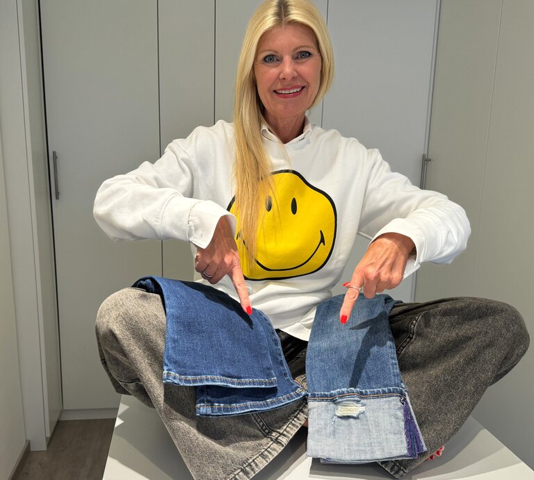 Renate Zott trägt lächelnd einen weißen Pullover mit einem großen, gelben Smiley-Gesicht darauf und zeigt auf zwei verschiedene Jeansstoff-Muster, die sie auf ihren Knien platziert hat. | © Renate Zott