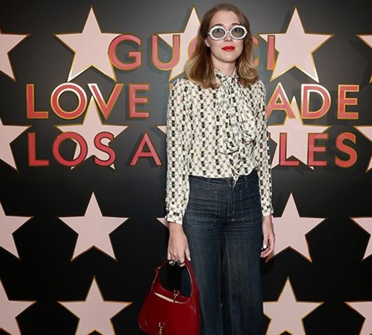Angelica Hicks vor einer Wand mit Sternen und einem Schriftzug. Sie trägt eine modische, gemusterte Bluse, eine dunkle Jeans und eine rote Handtasche, außerdem eine auffällige, runde Sonnenbrille. | © Getty Images /	Matt Winkelmeyer 