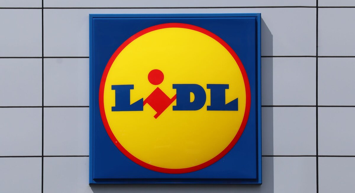  Das Foto zeigt das quadratische Firmenschild des Einzelhändlers Lidl. Das Schild ist auffallend gestaltet mit einem gelben Kreis auf blauem Hintergrund und dem Lidl-Logo in Blau und Rot in der Mitte. Es ist an einer Außenwand mit weißen, quadratischen Fliesen montiert.