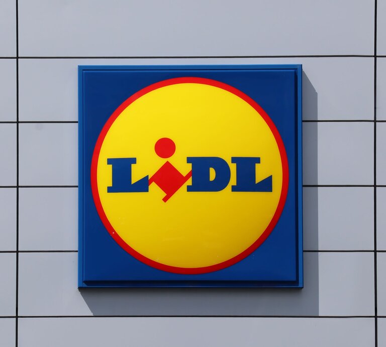  Das Foto zeigt das quadratische Firmenschild des Einzelhändlers Lidl. Das Schild ist auffallend gestaltet mit einem gelben Kreis auf blauem Hintergrund und dem Lidl-Logo in Blau und Rot in der Mitte. Es ist an einer Außenwand mit weißen, quadratischen Fliesen montiert. | © Adobe Stock/ Andrei Antipov