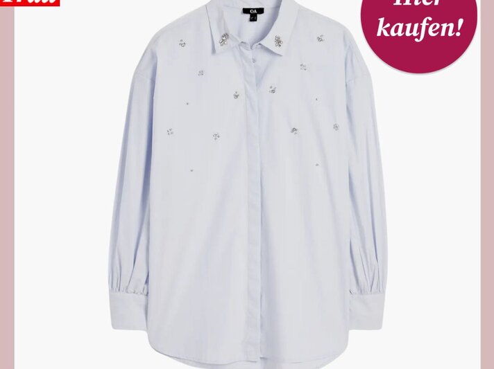 Ein Werbebild einer hellblauen Bluse mit dezentem, eingestreutem Blumenmuster. Die Bluse hat einen klassischen Kragen, lange Ärmel mit Manschetten und eine Knopfleiste auf der Vorderseite. Das Kleidungsstück ist flach und ordentlich ausgelegt, um das Muster und den Schnitt zu zeigen. Oben links im Bild befindet sich das Logo "Bild der Frau" in Weiß auf rotem Hintergrund, und oben rechts ist ein rundes, bordeauxrotes Etikett mit der weißen Aufschrift "Hier kaufen!". | © C&A