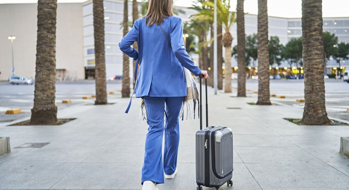 Frau in blauem Outfit von hinten mit Koffern, links und rechts Palmen