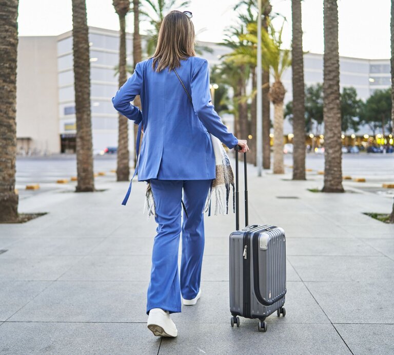Frau in blauem Outfit von hinten mit Koffern, links und rechts Palmen | © Getty Images