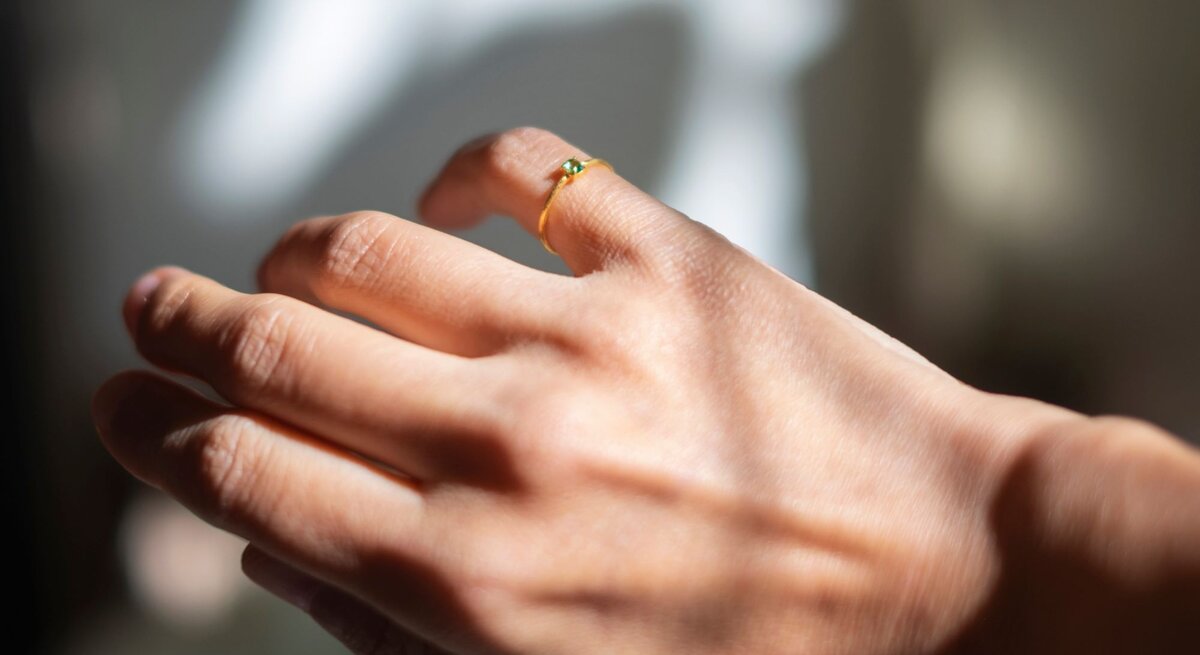 Eine Nahaufnahme einer Hand, die einen kleinen Ring mit einem grünen Stein am kleinen Finger trägt. Die Hand ist im Sonnenlicht, was für einen schönen Glanz und Schattenspiel sorgt. Der Hintergrund ist unscharf, was den Fokus auf den Ring und die Hand legt."