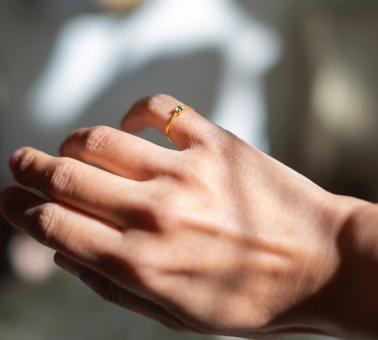 Eine Nahaufnahme einer Hand, die einen kleinen Ring mit einem grünen Stein am kleinen Finger trägt. Die Hand ist im Sonnenlicht, was für einen schönen Glanz und Schattenspiel sorgt. Der Hintergrund ist unscharf, was den Fokus auf den Ring und die Hand legt." | © Adobe Stock/ Nii Koo Nyan