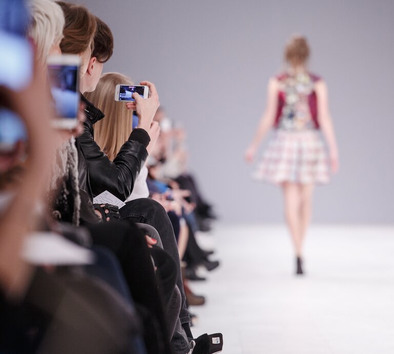Das Bild zeigt eine Modeschau aus der Perspektive des Publikums. Im Vordergrund sind unscharf Zuschauer zu sehen, von denen einige Fotos mit ihren Smartphones machen. Die Aufmerksamkeit ist auf das Model im Hintergrund gerichtet, das gerade den Laufsteg entlanggeht. Das Model ist von hinten zu sehen und trägt ein modisches Outfit, das aus einem Rock und einem Oberteil mit auffälligem Design besteht. Der Fokus und die Komposition des Fotos betonen die Dynamik und das Ambiente einer Fashion-Show, bei der das Erlebnis und das Festhalten des Moments im Vordergrund stehen. | © AdobeStock/oksanazahray