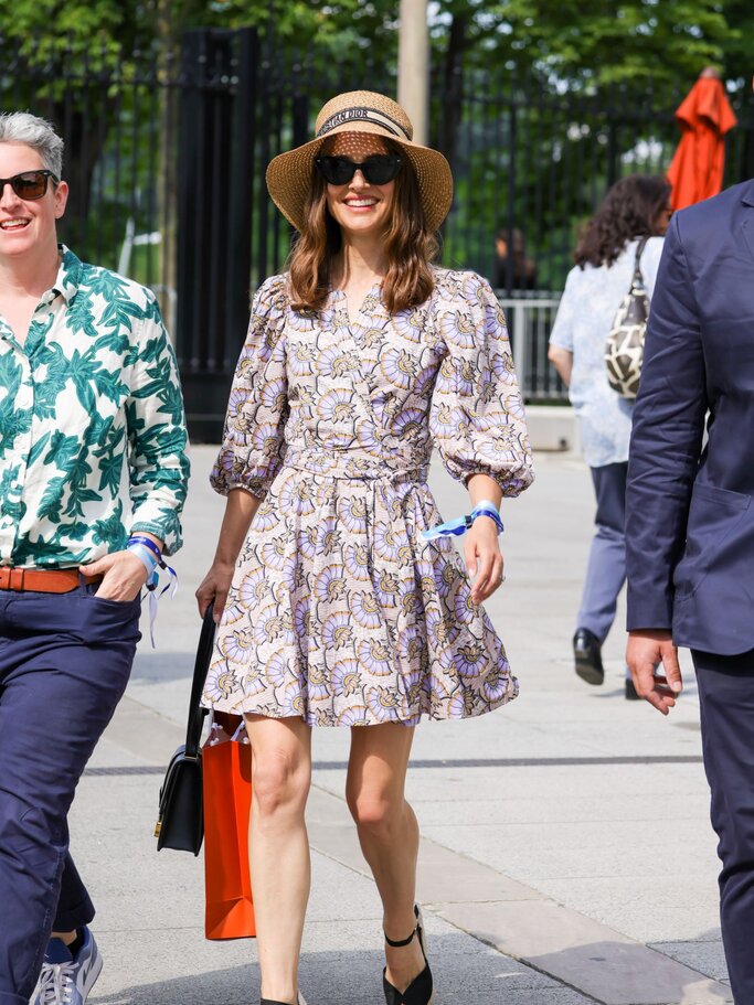 Natalie Portman mit Strohhut und geblümtem Sommerkleid läuft in Keilabsätzen durch eine sonnige Stadtstraße. | © Getty Images/ Pierre Suu / Kontributor