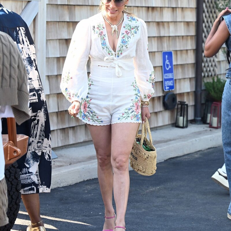 Jennifer Lopez mit strengem Pferdeschwanz trägt weißen bestickten Einteiler, rosa Keilsandaletten und Basttasche bei Sonnenschein. | © IMAGO / Cover-Images