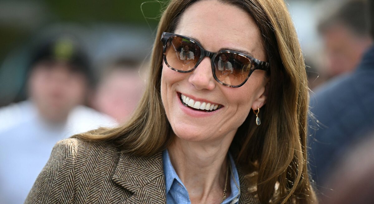 Prinzessin Kate mit glatten braunen Haaren, Sonnenbrille und Ohrhänger trägt braunen Blazer über Jeanshemd in sonniger Menschenmenge.