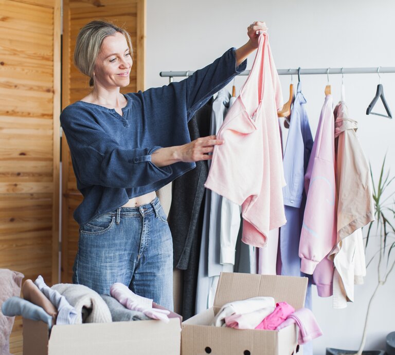 Eine blondhaarige Frau in Jeans und blauem Oberteil hält ein rosafarbenes Shirt vor Kleiderständer und Kartons mit Kleidung hoch. | © Adobe Stock/ Tatyana Gladskih