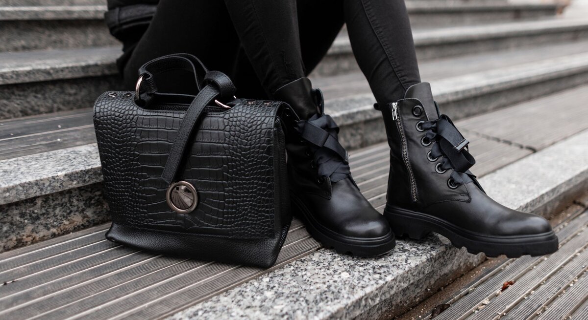 Nahaufnahme von schwarzen Lederstiefeln und einer luxuriösen Handtasche auf einer Treppe aus Stein und Holz.