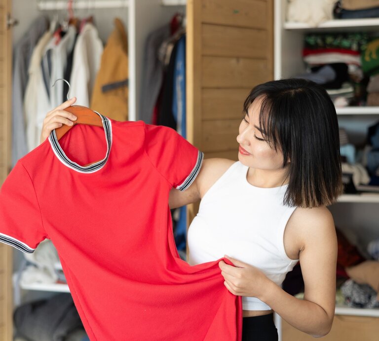 Frau, die ein rotes Oberteil in der Hand hält und es betrachtet. Im Hintergrund ist ein offener Kleiderschrank mit verschiedenen Kleidungsstücken zu sehen. | © Adobe Stock /  JackF
