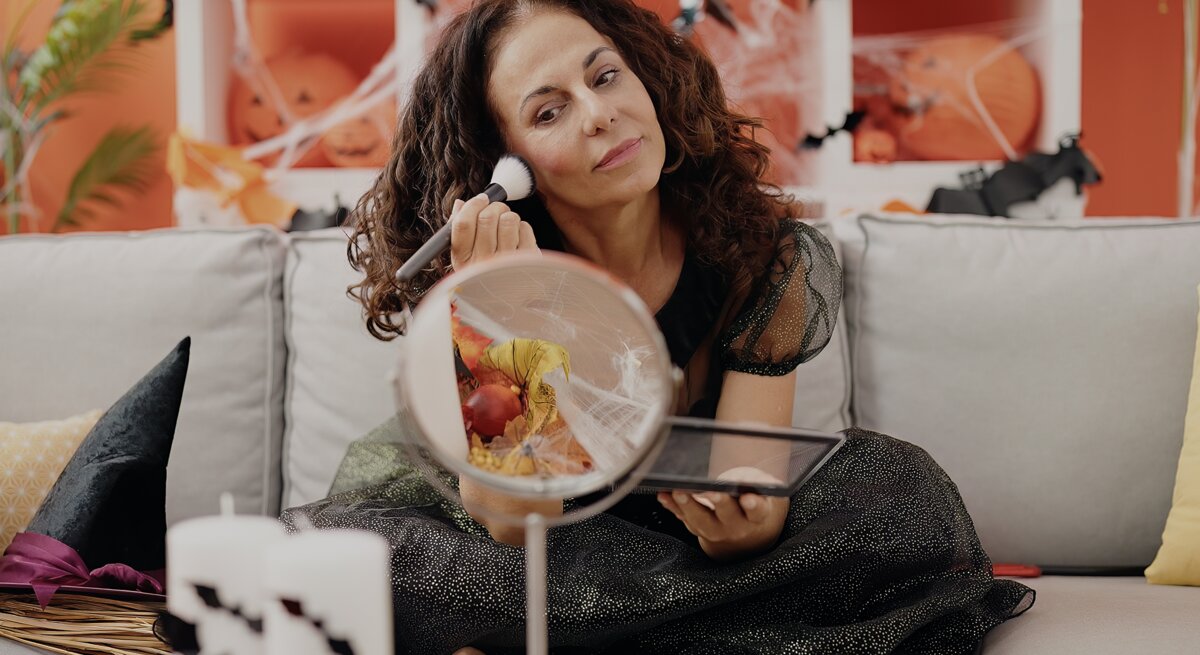 Eine Frau in glitzerndem schwarzen Kleid sitzt auf einem grauen Sofa, mit Halloween-Dekoration und Kürbissen im orangefarbenen Hintergrund.