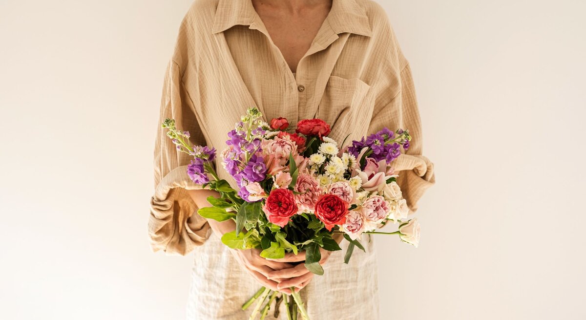 Frau mit kurzem braunem Haar hält einen bunten Strauß Blumen vor cremefarbenem Hintergrund und blickt zur Seite.