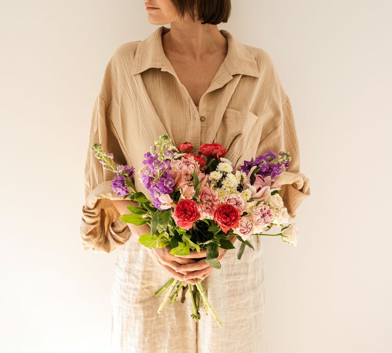Frau mit kurzem braunem Haar hält einen bunten Strauß Blumen vor cremefarbenem Hintergrund und blickt zur Seite. | © Adobe Stock/ Floral Deco