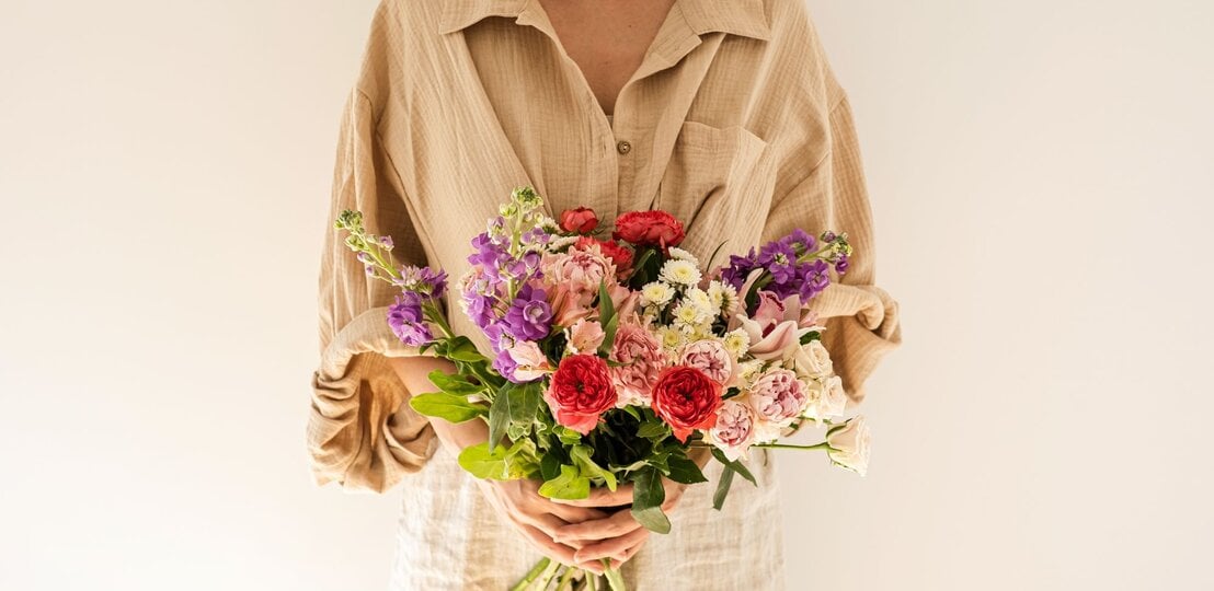 Frau mit kurzem braunem Haar hält einen bunten Strauß Blumen vor cremefarbenem Hintergrund und blickt zur Seite. | © Adobe Stock/ Floral Deco