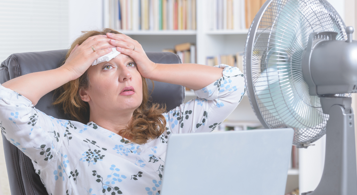 Eine mittelalte Frau mit braunen Haaren sitzt erschöpft vor Laptop und Ventilator im Büro mit Bücherregal im Hintergrund.