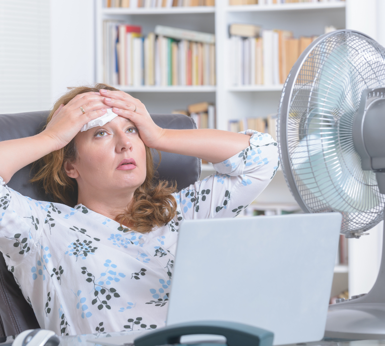 Eine mittelalte Frau mit braunen Haaren sitzt erschöpft vor Laptop und Ventilator im Büro mit Bücherregal im Hintergrund. | © Canva