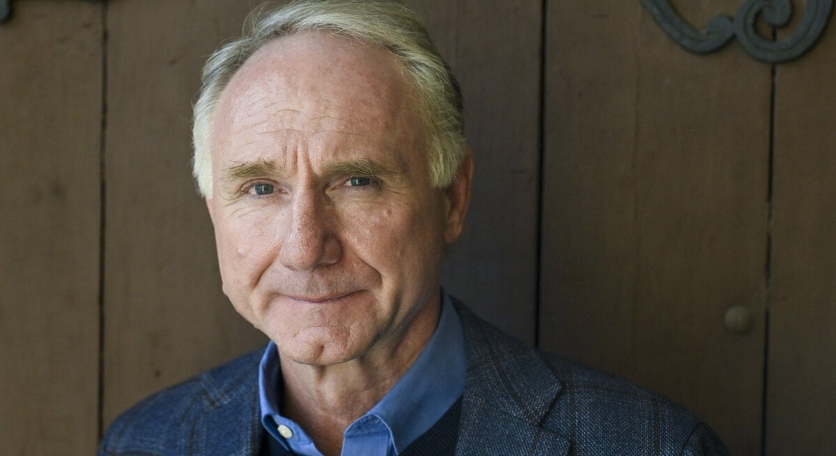 Dan Brown trägt ein dunkles kariertes Sakko mit blauem Hemd und Pullover, im Hintergrund eine schlichte Holzwand mit Metallverzierungen.
