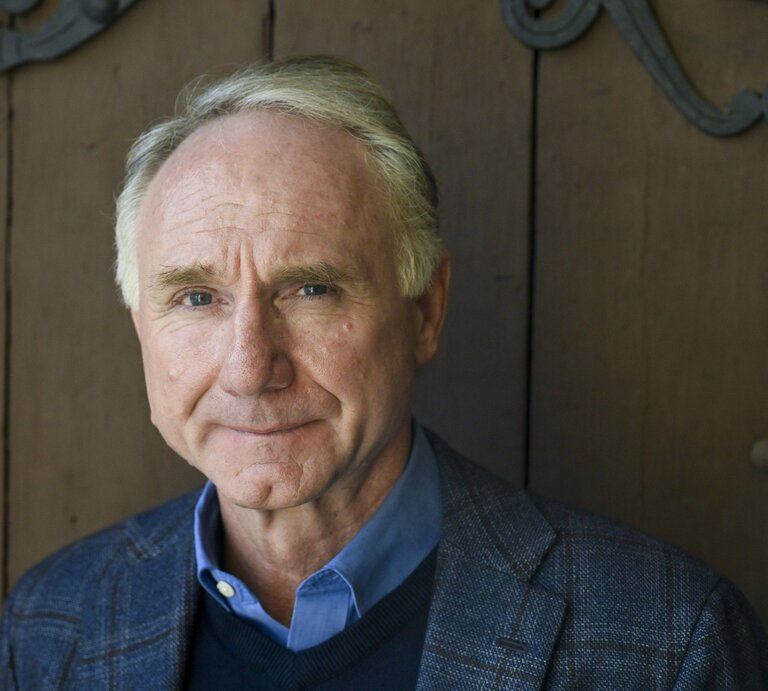 Dan Brown trägt ein dunkles kariertes Sakko mit blauem Hemd und Pullover, im Hintergrund eine schlichte Holzwand mit Metallverzierungen. | © IMAGO / Lehtikuva