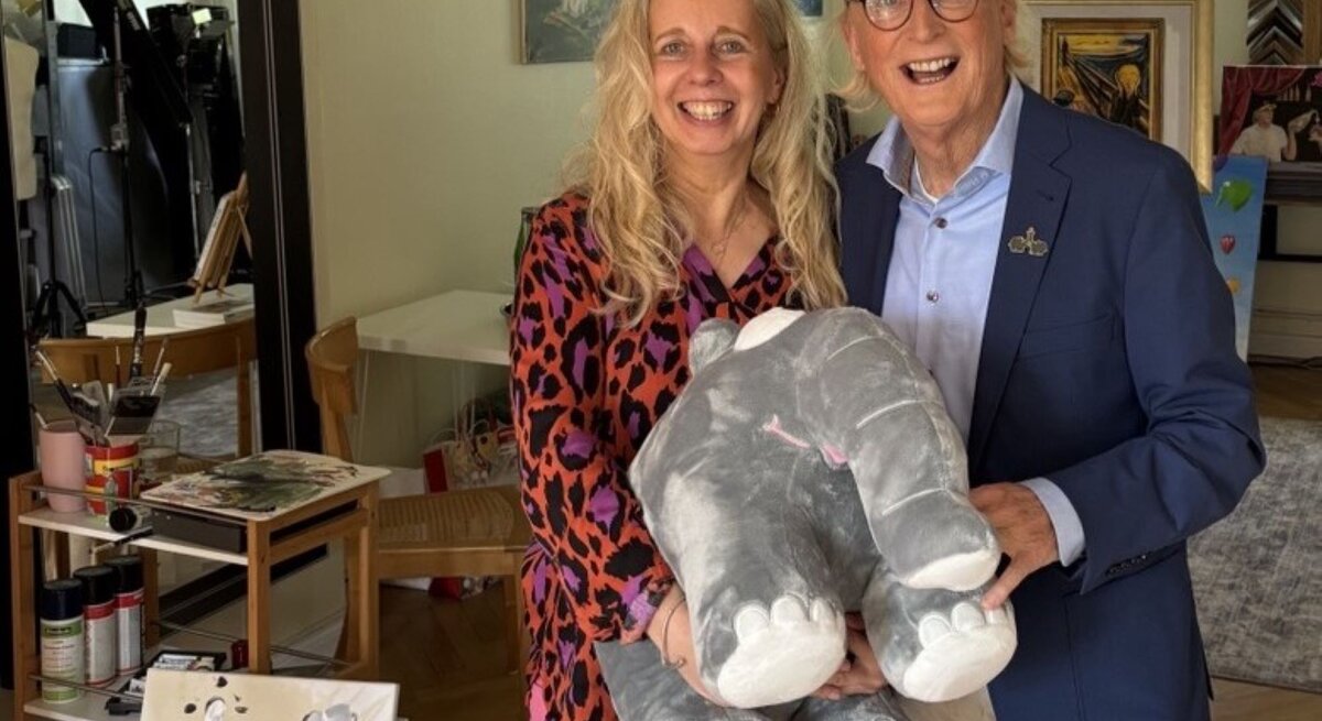 Eine blonde Frau in bunter Bluse und ein älterer Mann mit Brille und Basecap halten lachend ein graues Stoffelefanten-Kuscheltier.