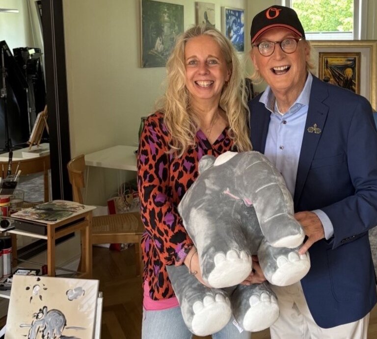 Eine blonde Frau in bunter Bluse und ein älterer Mann mit Brille und Basecap halten lachend ein graues Stoffelefanten-Kuscheltier. | © BILD der FRAU
