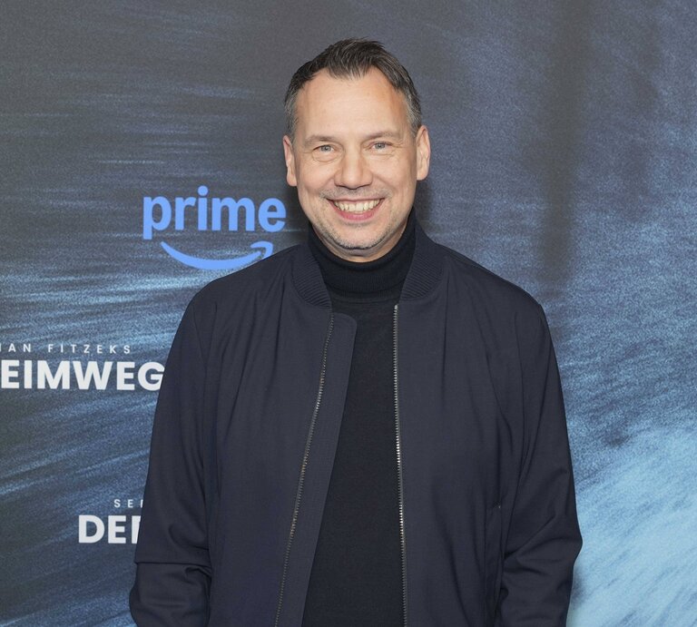 Sebastian Fitzek in dunkelblauer Jacke und schwarzem Rollkragenpullover steht vor einer Fotowand mit Schriftzug und Prime-Logo. | © IMAGO / BREUEL-BILD