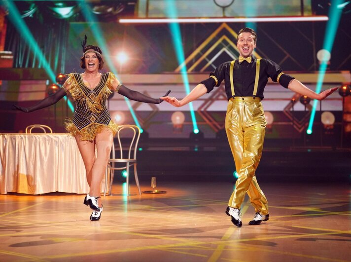 Lachendes Tanzpaar (Christine Neubauer & Valentin Lusin) im goldenen Charleston-Outfit tanzt synchron auf beleuchtetem Parkett vor Art-Déco-Kulisse. | © RTL / Stefan Gregorowius