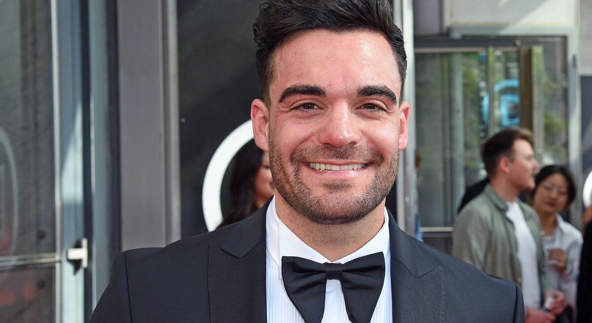 Stefano Zarrella: Ein junger Mann mit dunklen Haaren und Bart lächelt in einem schwarzen Smoking mit Fliege vor einer gläsernen Event-Location.